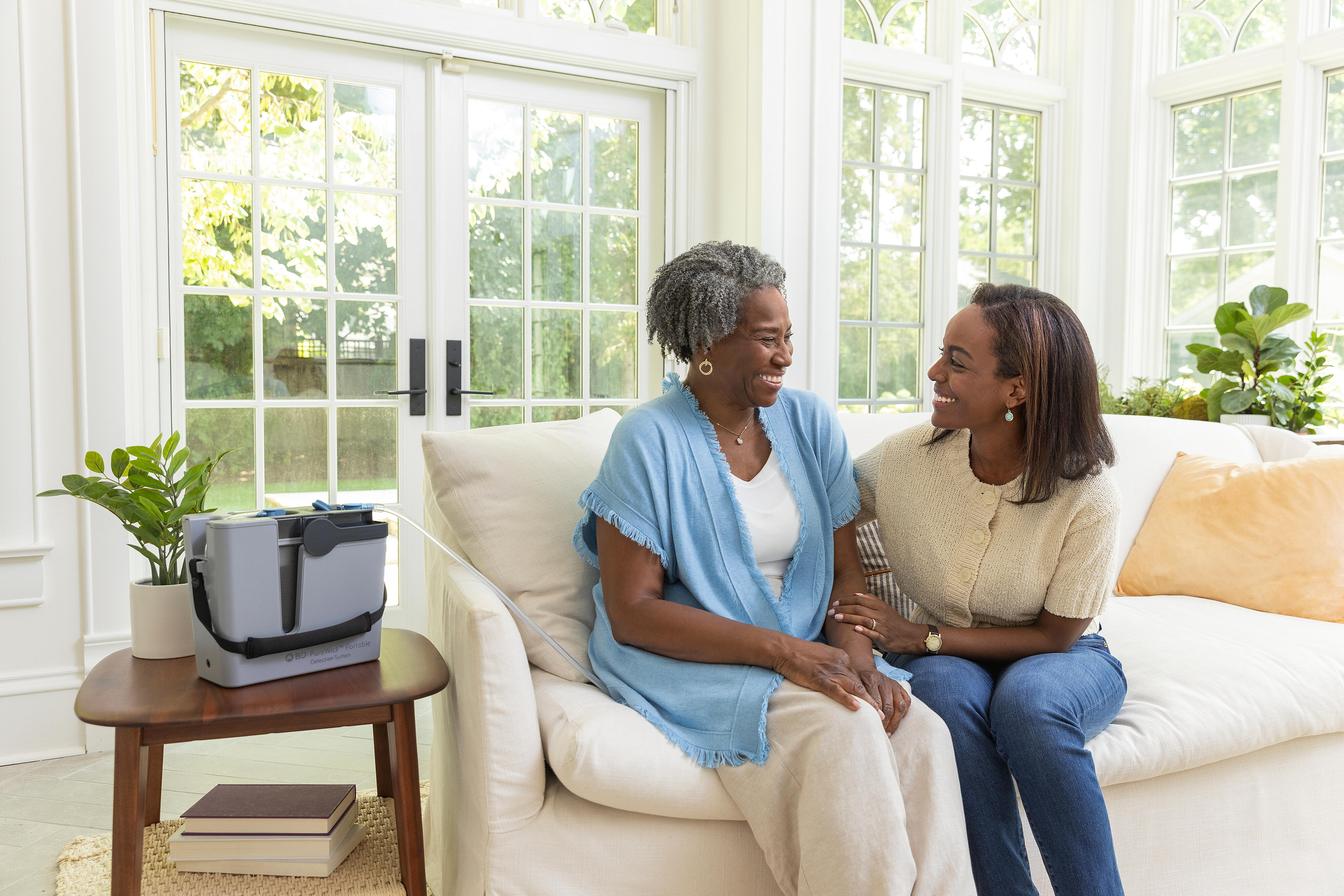 Mother and daughter smiling at each other on couch with PureWick&trade; Portable Collection System on table.t goes here