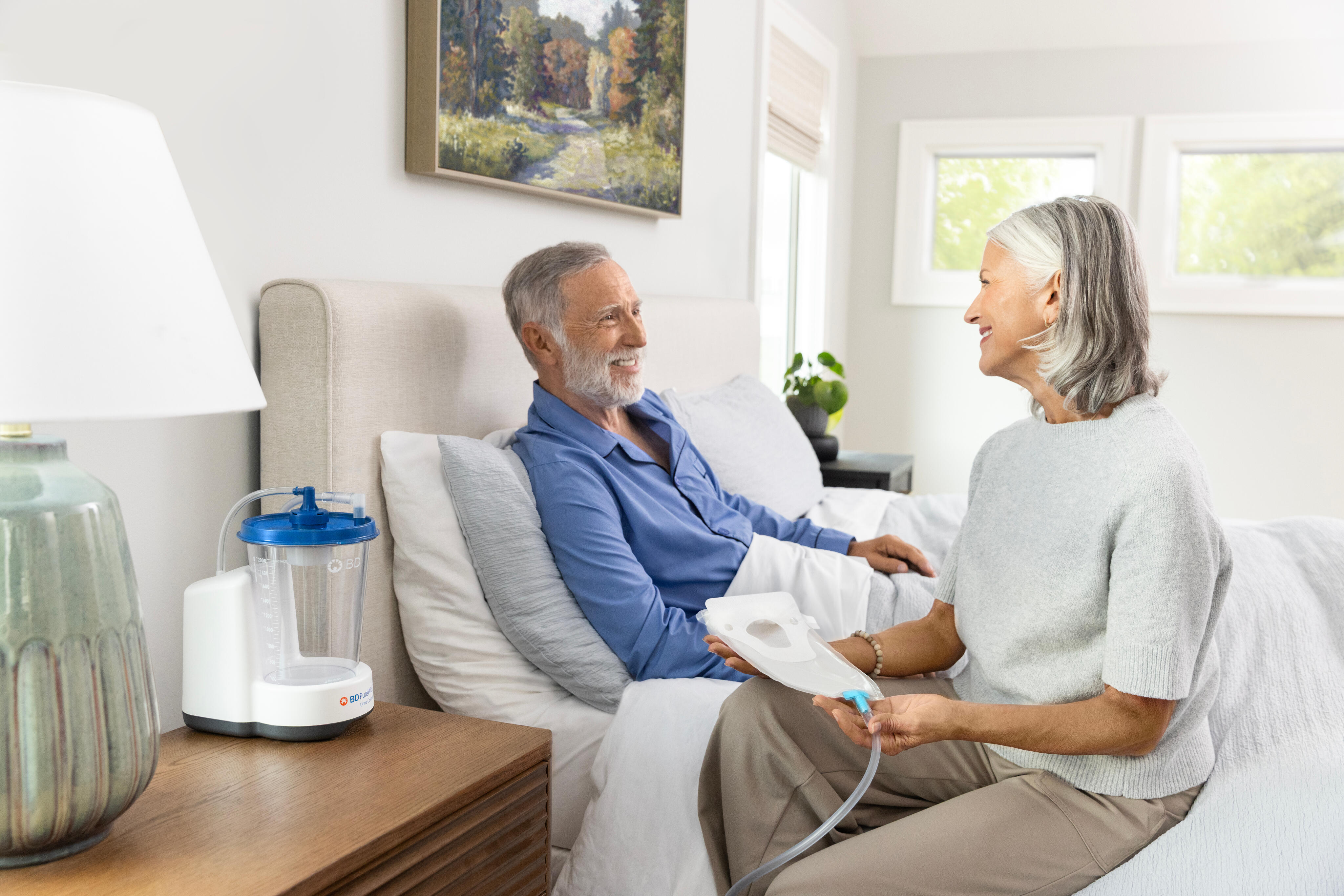 Woman and man smiling at each other in bed while she holds a PureWick&trade; External Catheter