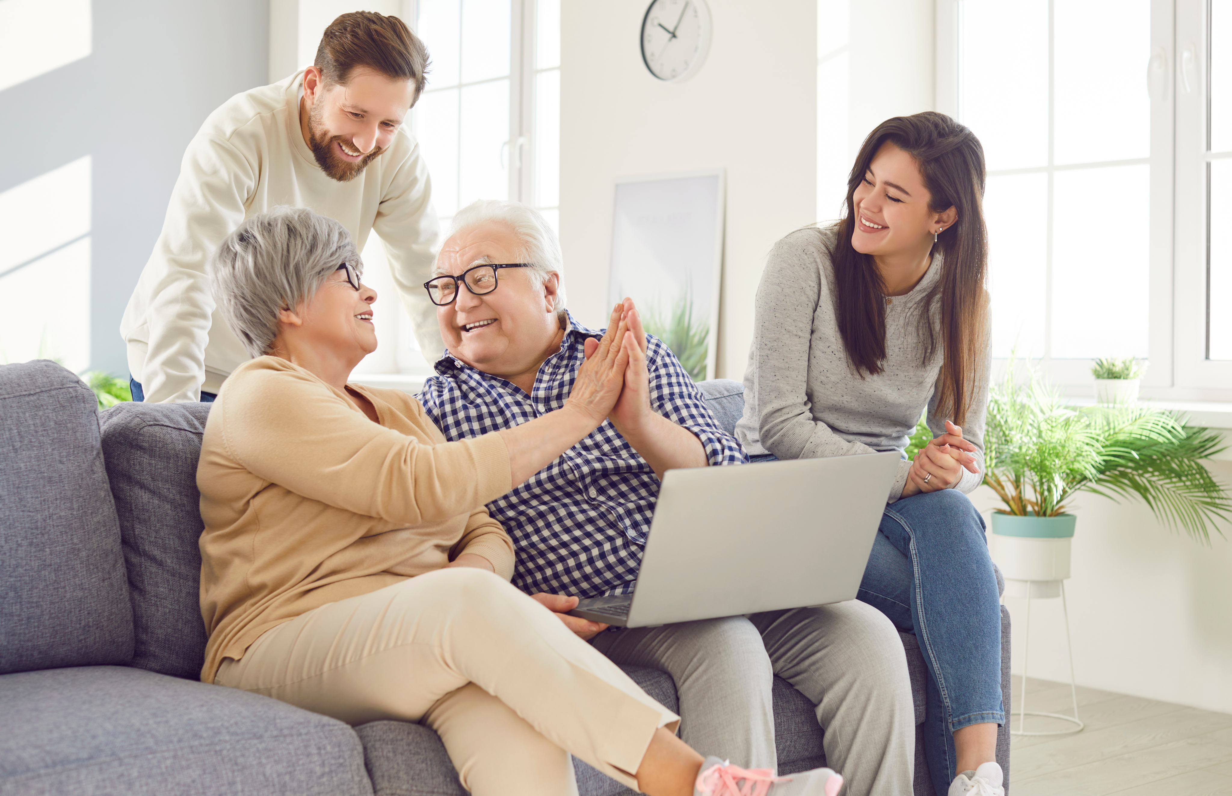 Elderly couple sitting on couch smiling and high-fiving each other while family members smile.